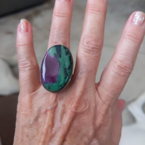 May include: A silver ring with a large oval green, pink, and purple gemstone. The ring is on a person's finger.