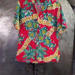 May include: Red short-sleeved button-up shirt with a tropical print of pineapples and palm trees.