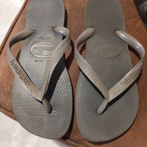 May include: A pair of gray flip-flops with the brand name "havaianas" printed on the straps. The flip-flops have a textured sole and a simple design.