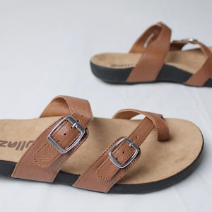 May include: Two brown leather sandals with adjustable straps and silver buckles. The sandals have a light brown footbed and a black sole. The word "zillaz" is printed on the footbed of one sandal. The sandals are displayed on a white surface.