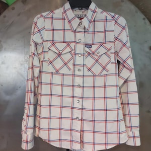 May include: White button-down shirt with a plaid pattern in red and blue. The shirt has two chest pockets and the brand name "DIXXON" is visible on the label and pocket. The shirt is hanging on a black hanger.