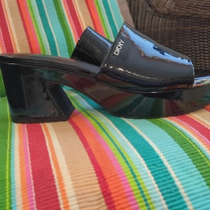 May include: Black platform sandals with a glossy finish. The sandals feature a chunky heel and a silver-toned trim around the platform. The brand name "DKNY" is visible on the strap. The sandals are placed on a colorful striped surface.