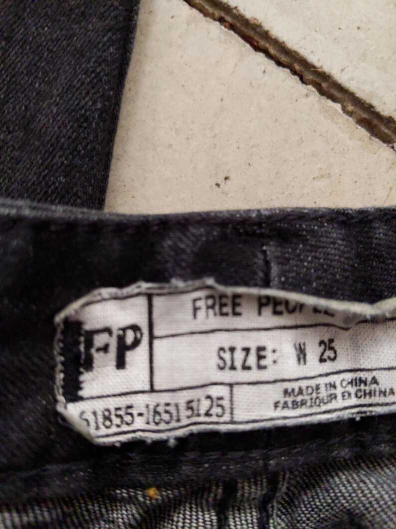 Puede incluir: Vaqueros de mezclilla negros con una etiqueta de Free People. La etiqueta tiene el texto "FP" y "SIZE: W 25" y los n&uacute;meros "$1855-1651 5125". La etiqueta tambi&eacute;n dice "MADE IN CHINA" y "FABRIQUER EN CHINA".