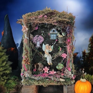 Op de afbeelding: Een grillig feeënhuis-diorama met een mos- en takkendak, met een miniatuur engel, bloemen en een vogelhuisje. Een oranje pompoen staat in de buurt, onder een maanverlichte hemel.