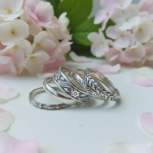 May include: A set of four stacked silver rings featuring various textures and patterns, including a thin band with fine detailing, a smooth polished band, a wider band with floral engravings, and a band with a repeating leaf-like pattern.
