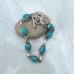 Puede incluir: Pulsera plateada con piedras turquesas ovaladas. La pulsera tiene un cierre de palanca y eslabones decorativos plateados. Las piedras turquesas son de un color azul-verdoso vibrante. La pulsera se muestra sobre una piedra gris lisa.