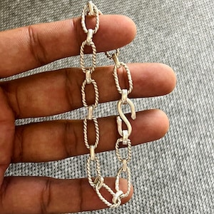 May include: Silver-toned chain bracelet featuring a series of oval and twisted links. The bracelet has an S-shaped clasp for closure. The design is simple and elegant, suitable for everyday wear.