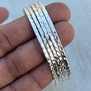 May include: A set of five silver bangles with a hammered texture. The bangles are stacked together, showcasing their reflective, shiny surface. The jewelry is held in a hand, with a soft, blurred background.