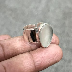 May include: A silver-toned ring with a large, teardrop-shaped, pale gemstone. The ring has a wide band and an open design, allowing for size adjustment. The gemstone is smooth and translucent, set in a polished metal setting.