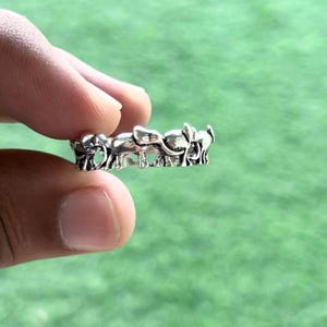 May include: A silver ring with a repeating elephant pattern. The ring is held between fingers against a blurred green background. The elephants are detailed with trunks, ears, and tails.