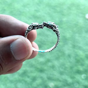 May include: A silver ring featuring two dragon heads facing each other. The ring has intricate detailing and a textured band. The dragons have detailed scales and facial features. The ring is held against a green background.