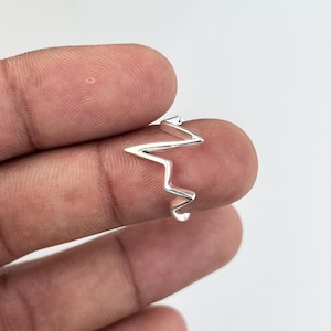 Può includere: Un anello color argento con un design a zig-zag. L'anello è tenuto tra due dita, mettendo in risalto la sua forma unica. Il design dell'anello ricorda un battito cardiaco o un'onda sonora, aggiungendo un tocco moderno al gioiello.