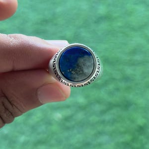 Könnte beinhalten: Ein silberner Ring mit einem großen, runden, blauen Edelstein in der Mitte. Der Edelstein hat ein marmoriertes Aussehen mit helleren grauen Flecken. Das Ringband weist ein sich wiederholendes geometrisches Muster auf.