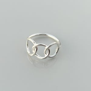 Może przedstawiać: Srebrny pierścionek z motywem połączonych ogniw łańcucha. Pierścionek ma trzy połączone owalne kształty, tworzące ciągłą pętlę. Pierścionek jest wykonany z błyszczącego, odbijającego światło metalu, na neutralnym tle.