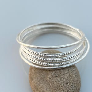 May include: A stack of silver rings, some with a smooth finish and others with a dotted texture. The rings are resting on a small, light brown stone, with a soft, light blue background.