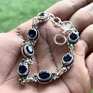 Puede incluir: Pulsera de plata con piedras preciosas azules ovaladas y redondas. La pulsera tiene un diseño ornamentado con cierre de broche. Las piedras preciosas están engastadas en una montura de metal plateado.