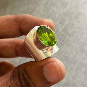 Pode incluir: Um anel de prata com uma grande pedra preciosa verde brilhante de corte oval. A pedra preciosa é engastada numa banda de prata polida com um design angular único. O anel é segurado contra um fundo cinzento neutro.