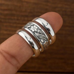 Puede incluir: Un conjunto de cuatro anillos de plata. Uno presenta un diseño floral con hojas, mientras que los otros tres son bandas lisas. Los anillos están apilados en un dedo, mostrando sus superficies pulidas y reflectantes. El fondo es una superficie de madera borrosa.
