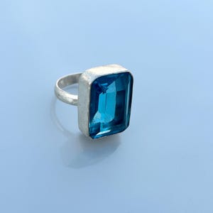 Könnte beinhalten: Ein silberner Ring mit einem großen, rechteckigen, leuchtend blauen Edelstein. Der Edelstein ist in eine silberne Lünette gefasst, und der Ringband ist ebenfalls silberfarben. Der Ring wird auf einer hellblauen Oberfläche gezeigt.
