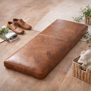May include: A rectangular, brown leather ottoman with a stitched border and a subtle logo. The ottoman is placed on a wooden floor, with a pair of brown leather shoes and a magazine nearby. A woven basket and plants are also in the scene.
