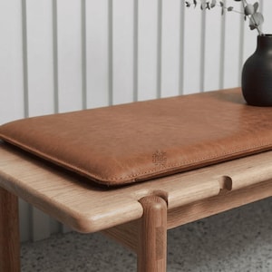 May include: A rectangular tan leather bench cushion with visible stitching along the edges and a small embossed logo detail. The cushion is placed on a light-coloured wooden bench, providing a smooth, flat surface for seating comfort.