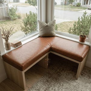 May include: An L-shaped corner bench with a light wood base and a smooth, brown faux leather seat cushion. A single square, textured beige throw pillow rests on the bench. A small ceramic vase with dried flowers and two stacked books sit nearby.