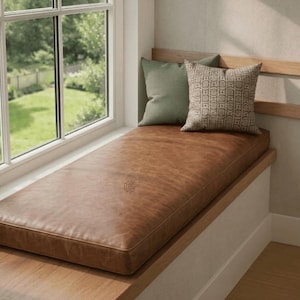 May include: A rectangular brown leather window seat cushion featuring a smooth surface and piped edges. Two square throw pillows are placed on top: one solid sage green pillow and one patterned pillow with a geometric brown and beige design.