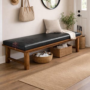 May include: A wooden entryway bench with a black leather cushion. A woven basket and a beige rug are in front of the bench. A round mirror and a coat rack with a hat and tote bag are on the wall.