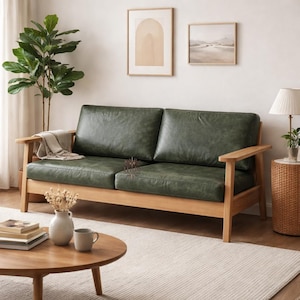 May include: A two-seater sofa with a green leather seat and backrest, set on a light-coloured wooden frame. The sofa is in a living room setting, with a round wooden coffee table, a rug, and framed artwork on the wall.