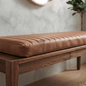 May include: A rectangular bench featuring a dark brown wooden frame and a tan leather seat cushion with vertical channel tufting. The cushion surface displays a small, centered logo with the text Hide and a stylized tree graphic.
