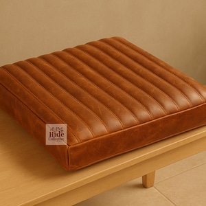 May include: A square, brown leather cushion with vertical stitched lines. The cushion is placed on a light-colored wooden bench. The cushion has a rectangular shape and is made of a soft, textured material.