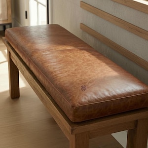 May include: A rectangular bench with a brown leather cushion and a wooden frame. The leather has a textured appearance and a small decorative emblem is visible on the cushion. The wooden frame has a natural finish.