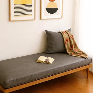 May include: A gray upholstered bench with a wooden frame, a gray pillow, and a floral throw. Two framed abstract art prints hang on the wall. An open book rests on the bench. The room has hardwood floors.