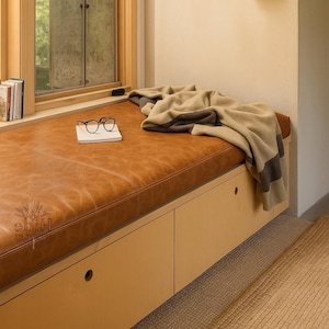 May include: A built-in window seat with a brown leather cushion, a folded blanket, and a small book with glasses. The seat has light wood cabinetry below. A window is visible in the background.
