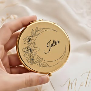May include: A gold-toned, circular compact mirror featuring a floral and crescent moon motif. The mirror is engraved with the name "Julia" in cursive script. The floral design includes lilies and foliage.