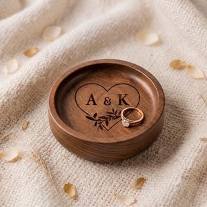 Pode incluir: Um prato redondo de madeira castanho-escura com um design de coração gravado a laser, as letras A & K e um pequeno ramo com folhas. Um anel fino de ouro rosa com uma pedra transparente repousa dentro da taça de madeira.