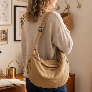 May include: A tan canvas hobo bag with a wide adjustable shoulder strap and a curved silhouette. The bag features a prominent horizontal zipper closure across the top and a secondary zipper compartment on the front, crafted from durable fabric.
