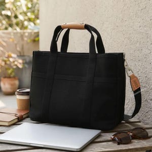 Puede incluir: Un bolso tote negro con un asa de cuero marrón claro y una correa de hombro negra desmontable. El bolso tiene dos asas cortas y varios bolsillos. Una computadora portátil, gafas de sol y una taza de café están sobre la mesa de madera.