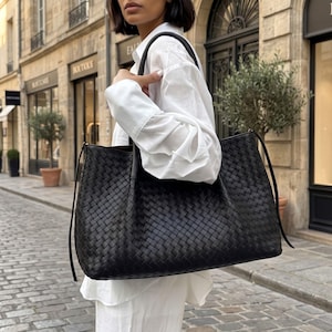 May include: A large black tote bag featuring an intricate woven leather pattern. The bag has two thin shoulder straps and side ties, showcasing a structured rectangular shape with a matte finish and a consistent grid-like texture throughout.