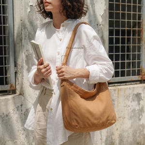May include: A tan suede shoulder bag with a curved design. The bag has a long strap and a large main compartment. The word "Personalization" is printed on the strap. The bag is worn over the shoulder.