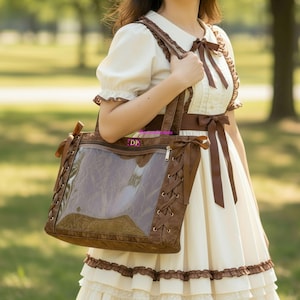 May include: A brown tote bag with a clear front panel and brown trim. The bag has brown handles and decorative lacing on the sides. The bag is being carried by a person wearing a cream-coloured dress with brown accents.