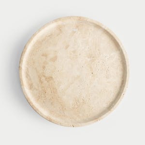 Puede incluir: Una bandeja redonda de mármol beige con un borde ligeramente elevado. La bandeja tiene una superficie natural y texturizada con sutiles variaciones de color, mostrando los patrones únicos de la piedra. Ideal para servir o exhibir.