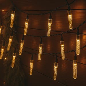 Puede incluir: Cadena de luces LED blanco cálido con bombillas cilíndricas transparentes. Las luces están ensartadas en un cable verde oscuro sobre un fondo de madera. Las bombillas tienen un aspecto texturizado y burbujeante, y están encendidas.