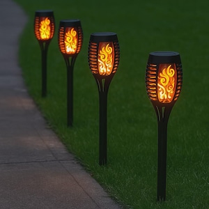 May include: Four black solar-powered torches with a flame effect are arranged in a row along a walkway and grass. The torches have a decorative cage design around the flame-simulating LED lights, creating a warm, inviting ambiance for outdoor spaces.