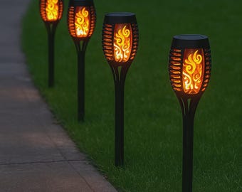 May include: Four black solar-powered torches with a flame effect are arranged in a row along a walkway and grass. The torches have a decorative cage design around the flame-simulating LED lights, creating a warm, inviting ambiance for outdoor spaces.