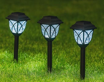 Könnte beinhalten: Drei schwarze Solar-Gartenleuchten mit dekorativem Glasdesign. Die Leuchten strahlen ein sanftes weißes Licht aus und stehen auf einem grünen Rasen. Jede Leuchte hat eine klassische Laternenform.