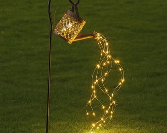 Könnte beinhalten: Gartenstecker mit einem dekorativen Gießkannen-Design. Die Gießkanne aus Metall hat ein Gittermuster und ist mit warmweißen LED-Lichtern beleuchtet, die wie Wasser nach unten fließen. Der Stecker steht in einer Rasenfläche.