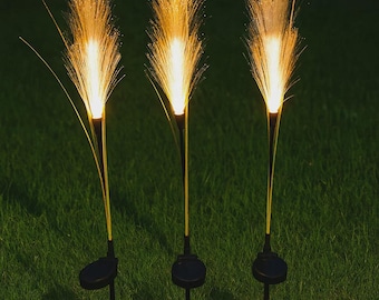 May include: Three solar-powered garden lights with a unique design. Each light features a black base and stem, topped with a cluster of illuminated, fiber-optic strands that resemble fireworks. The lights are set against a backdrop of green grass.