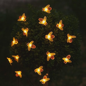 May include: A string of bee-shaped fairy lights. Each bee has a yellow body and wings, with a warm, glowing light. The lights are strung on a thin, dark wire, and are displayed against a backdrop of green foliage.