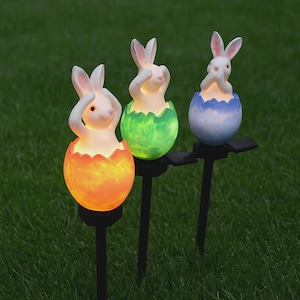 Puede incluir: Tres estacas de jardín con conejitos de Pascua. Cada conejito es blanco con orejas rosas y sale de un huevo colorido. Los huevos son naranjas, verdes y azules, y las estacas son negras. Las luces son solares.